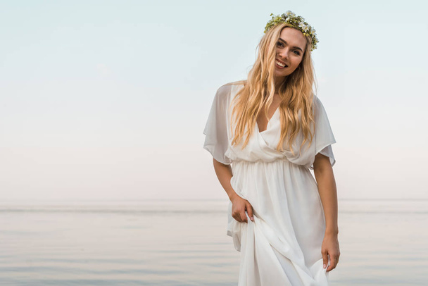 sonriente novia atractiva en vestido blanco y corona de flores mirando a la cámara cerca del océano
 - Foto, imagen