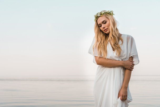 attractive bride in white dress and wreath of flowers hugging herself near ocean - Φωτογραφία, εικόνα