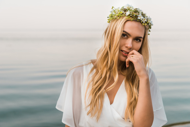 attractive bride in white dress and wreath of flowers touching lips and looking at camera on beach - Фото, изображение