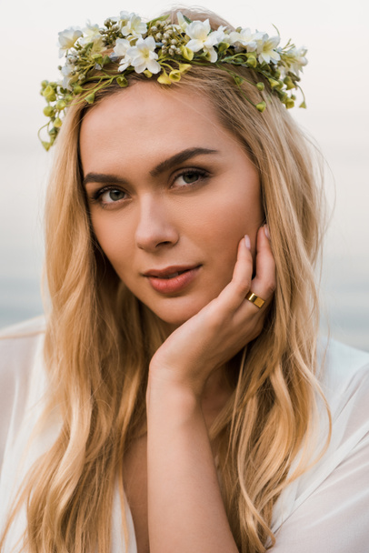 portrait of attractive bride in white dress and wreath of flowers looking at camera near sea - Photo, Image