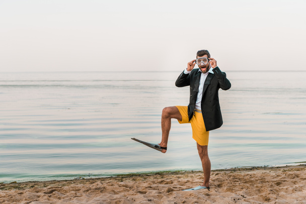 έκπληκτος ο άνθρωπος σε μαύρο σακάκι, σορτς και βατραχοπέδιλα αγγίζοντας μάσκα κολύμπι στην παραλία κοντά στη θάλασσα - Φωτογραφία, εικόνα