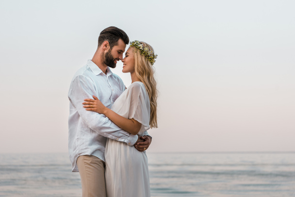 side view of bride and groom hugging and touching with noses on beach - Фото, зображення