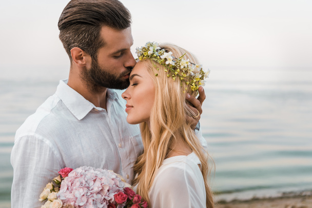 Seitenansicht des hübschen Bräutigams küsst attraktive Braut Stirn am Strand - Foto, Bild