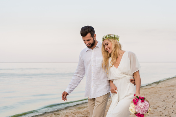 handsome groom hugging beautiful bride with wedding bouquet and they walking on beach - Photo, Image