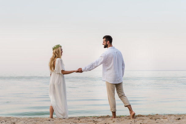 zadní pohled na nevěstu a ženicha, drželi se za ruce a v blízkosti oceánu při pohledu na sebe - Fotografie, Obrázek