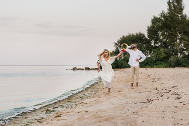 happy bride running with wedding bouquet, groom grimacing on beach - Foto, afbeelding