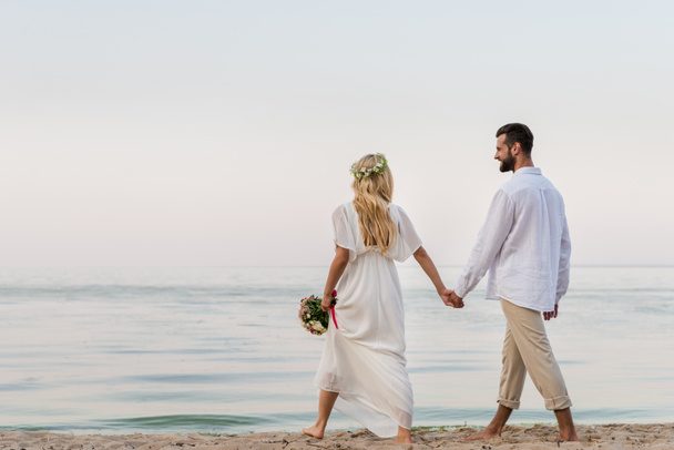 Arkadan gelin ve el ele tutuşarak ve düğün buket ile kumsalda yürüyen damat görünüşü - Fotoğraf, Görsel