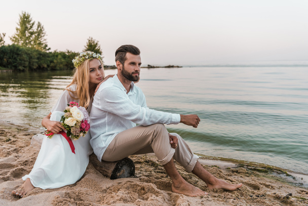 novio guapo y novia con ramo de bodas sentado en el registro en la playa
  - Foto, imagen