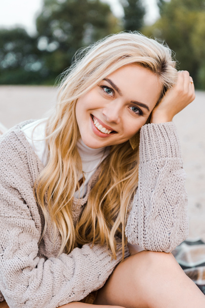 attractive smiling blonde girl in autumn outfit sitting on beach and looking at camera - Фото, зображення