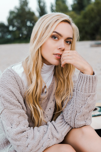attractive blonde woman in autumn outfit sitting on beach, touching lips and looking at camera - Фото, изображение