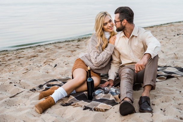 stylish couple in autumn outfit looking at each other on beach with bottle of red wine - Photo, Image
