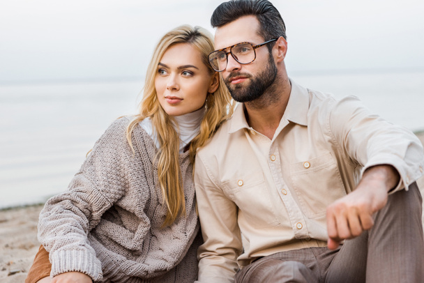 stylish couple in autumn outfit sitting on beach and looking away - Fotó, kép