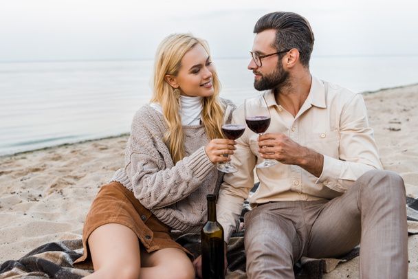 happy boyfriend and girlfriend in autumn outfit clinking with glasses on beach - Foto, afbeelding