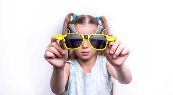 A smiling child girl in bright yellow sunglasses in the shape of a giraffe on a white background. - Zdjęcie, obraz