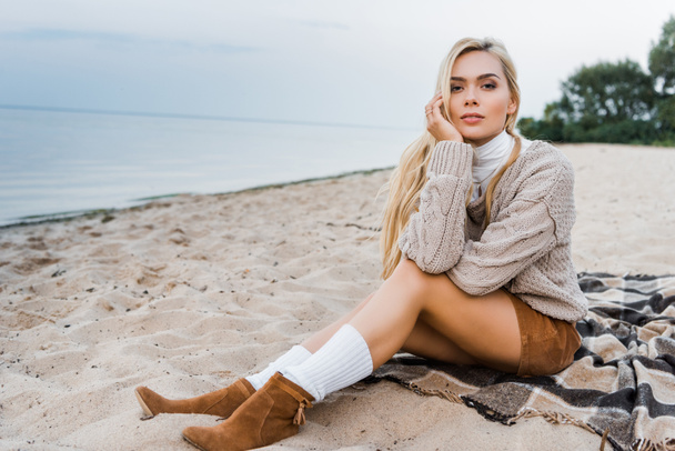 attractive blonde girl in autumn outfit sitting on beach, resting chin on hand and looking at camera - Photo, Image