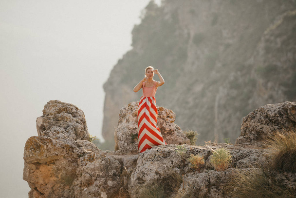 Привлекательная блондинка в красном платье позирует на вершине скалы с горой на заднем плане. Вид на горы. Вечер в Испании
 - Фото, изображение