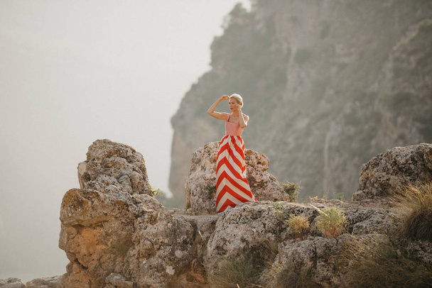 背景に山と崖の上に踊る赤いドレスの魅力的な金髪女性。マウンテン ビュー。スペインの夜 - 写真・画像