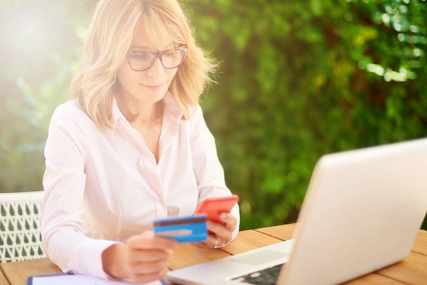 High angle shot of attractive middle aged woman shopping with credit card and using laptop while sitting in the garden at home.  - Foto, Imagen