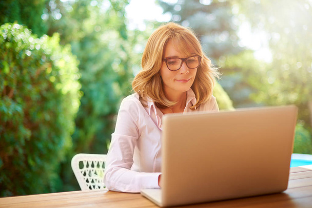 Confident middle aged woman sitting in the garden at home and using her laptop. Home office. - Photo, image