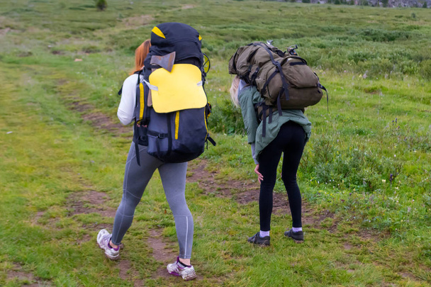 Twee meisjes toeristen met rugzakken gaan op de weg bedekt met stenen, het vermijden van een plas, de bergen en avonturen onder de grijze wolken, is een van hen moe met gestopt voor een rust - Foto, afbeelding