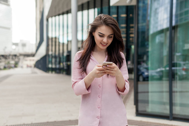 Smiling beautiful woman walking along the street using smartphone and doing online shopping, happy girl passing by a clothes shop window while browsing internet on her mobile phone and buying - Foto, imagen