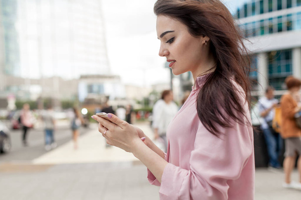 Happy pretty brunette girl or businesswoman using mobile phone, beautiful woman browsing phone and doing online shopping in a mobile app while smiling walking near shopping mall. Online communication - Фото, изображение