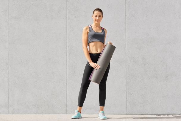 Photo of self determined female bodybuilder dressed in casual top, leggings and sportshoes, holds mat in hands, going to have exercises, isolated over concrete wall. People, sport, motivation concept - Photo, Image