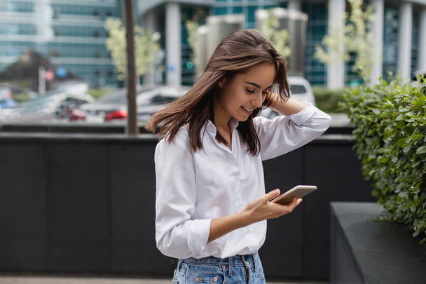 Happy pretty brunette girl business woman using mobile phone, beautiful woman browsing phone and doing online shopping in a mobile app while smiling walking near shopping mall. Online communication. - Φωτογραφία, εικόνα