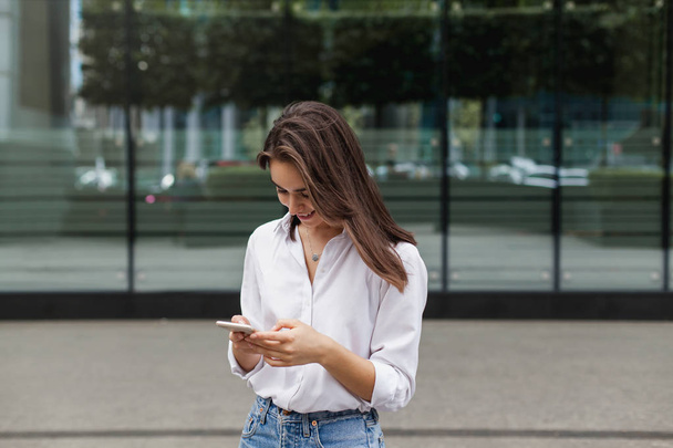 Happy pretty brunette girl using mobile phone near office, beautiful woman browsing phone and doing online shopping in a mobile app while smiling walking near shopping mall. Online communication. - Фото, зображення