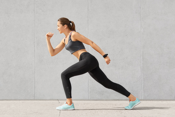 Horizontal shot of flexible woman with sporty body, stands in pose, ready to start marathon, dressed in sportsclothes, isolated over grey concrete wall. People. sport, determination concept. - Photo, image
