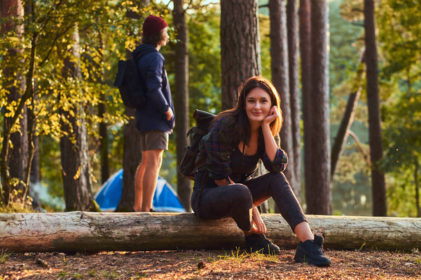 Nachdenkliches Touristenmädchen hält Hand aufs Kinn, während es auf einem Baumstamm sitzt und im Wald ein Smartphone in der Hand hält. - Foto, Bild