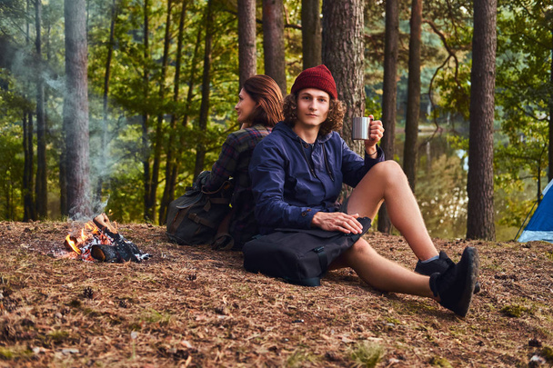 Fiatal turista házaspár ült vissza, vissza, miközben felmelegedés a következő látnivaló közelében: egy tábortűz a tábor az erdő. - Fotó, kép