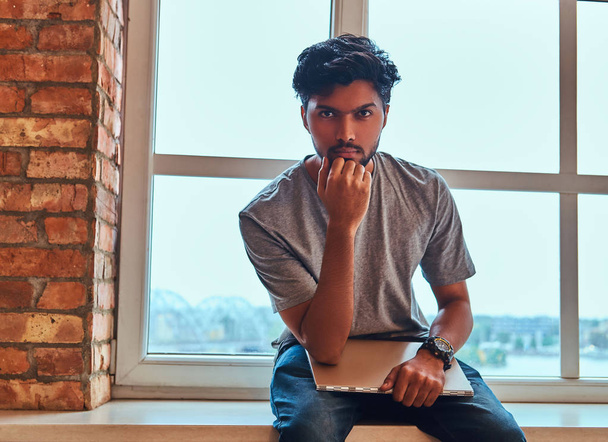 Zamyšlený indický student drží notebook zatímco sedí na okenním parapetu v kolejích. - Fotografie, Obrázek