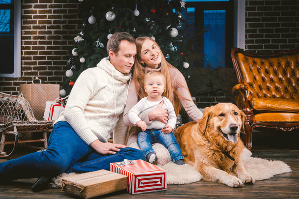 theme Christmas and New Year family circle and domestic pet. Mom dad and child 1 year old Caucasian woman sitting on floor near Christmas tree with gifts and big dog breed labrador golden retriever. - Fotoğraf, Görsel