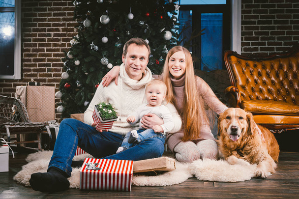 theme Christmas and New Year family circle and domestic pet. Mom dad and child 1 year old Caucasian woman sitting on floor near Christmas tree with gifts and big dog breed labrador golden retriever. - Photo, Image