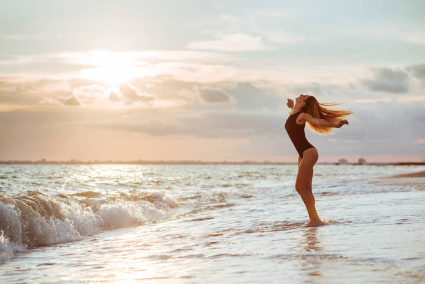Outdoor lifestyle portrait of beautiful girl in black swimsuit. Young hipster woman play with her hair. She is happy. Sunny hot summer day at tropical ocean beach. - 写真・画像