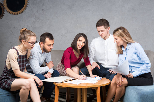 Subject business situation, teamwork. Group of young Caucasian five people sitting in office at a round table meeting, using paper, graphics. - Photo, Image