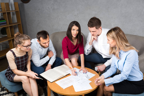 Sujet situation d'affaires, travail d'équipe. Groupe de jeunes Caucasiens cinq personnes assises au bureau lors d'une table ronde, en utilisant le papier, graphiques
. - Photo, image