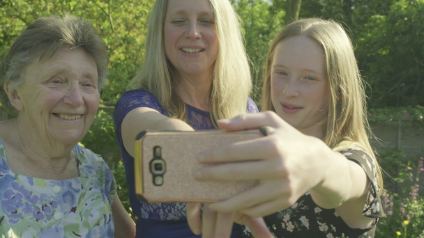 Isoäiti, hänen tyttärensä ja tyttärentyttärensä ottavat selfien älypuhelimella puutarhan ulkopuolella
 - Materiaali, video