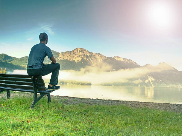 L'homme s'assied sur un banc en bois sur la côte du lac pittoresque ci-dessous sommets de montagne bleu. Calme, silence et harmonie concept
. - Photo, image