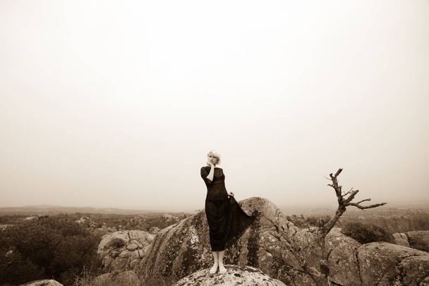 Foto artistica di una ragazza bionda in abito nero ed estetica sinistra sulle rocce in campagna in una giornata nuvolosa
.  - Foto, immagini