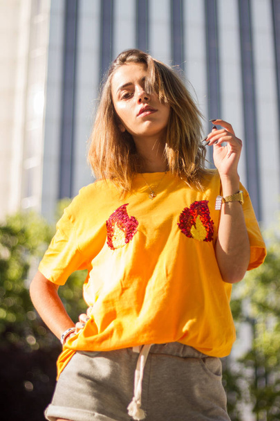 Portret van een jonge vrouw die zich voordeed op de straat, het dragen van een geel shirt terwijl de zon schijnt.  - Foto, afbeelding