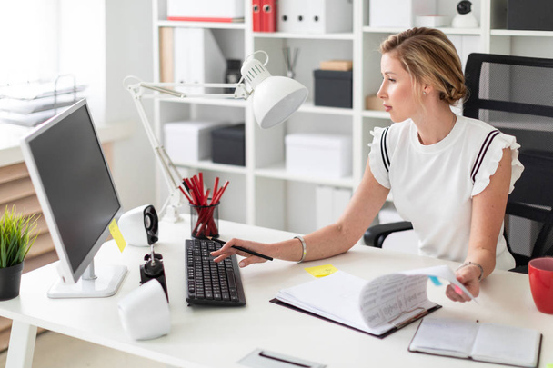 Ein junges blondes Mädchen sitzt an einem Computertisch im Büro, hält einen Bleistift in der Hand und arbeitet mit Dokumenten. - Foto, Bild