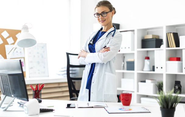 A young girl in a white coat is standing near a table in her office. A stethoscope hangs around her neck. - Foto, Bild