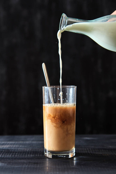 Coffee with milk. On a black wooden table, milk is poured into a glass of coffee. - Photo, image