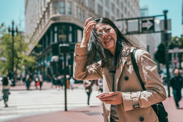 nuori aasialainen nainen seisoo kadulla Morden kaupungin ja etsivät onnellisesti ja pitää matkapuhelin
. - Valokuva, kuva