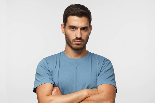 Young man in blue t-shirts tanding with arms crossed and serious concentrated face at camera, looking aggressive, isolated on gray background  - Foto, immagini
