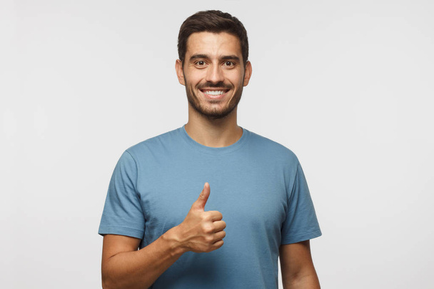 Young man isolated on grey background showing thumb up with positive emotions and happiness. Satisfaction with quality and recommendation - 写真・画像