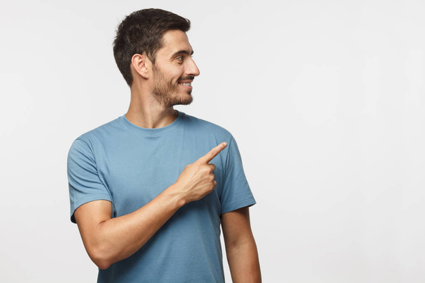 Casual dressed young man in blue t-shirt looking right, pointing with his finger, isolated on gray background with copy space for your text - Valokuva, kuva