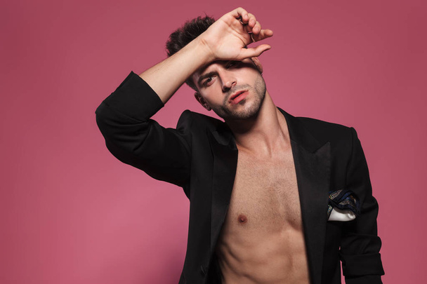 sensual man with naked chest and black undone tuxedo puts his hand on forehead, posing seductively while standing on red background, portrait picture - Photo, Image